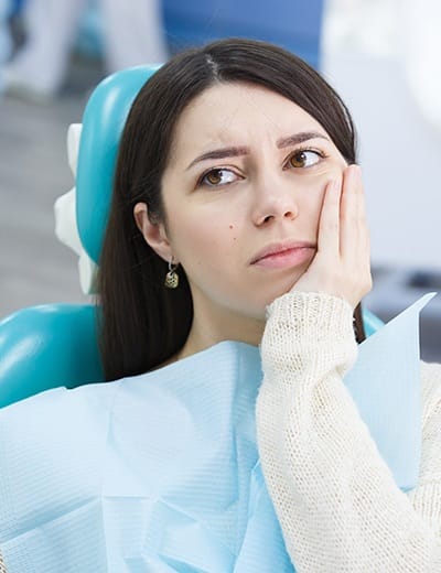 Woman in dental chair holding her cheek and wincing in pain