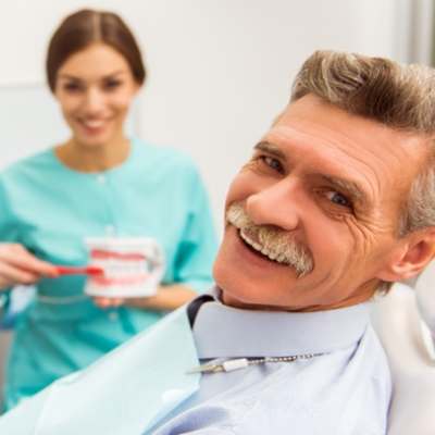 Man in dental chair smiling as dentist demonstrates how to brush dentures
